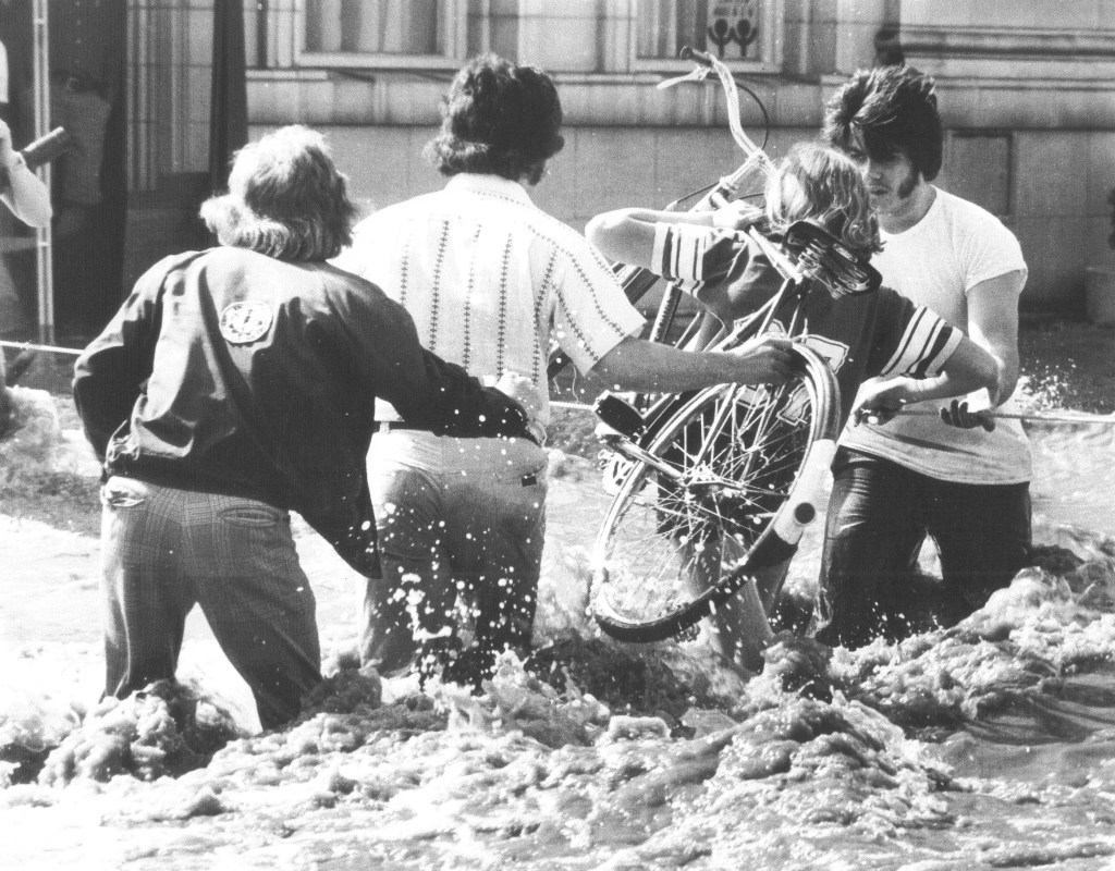 People carry a bicycle through choppy waters on a flooded city street.
