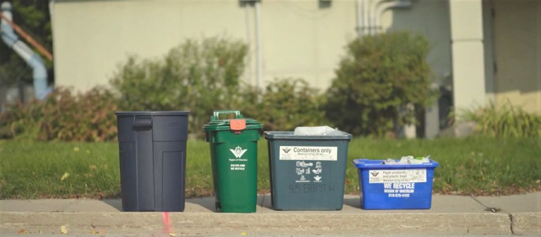A green bin, garbage can and blue box sit on a sidewalk curb.