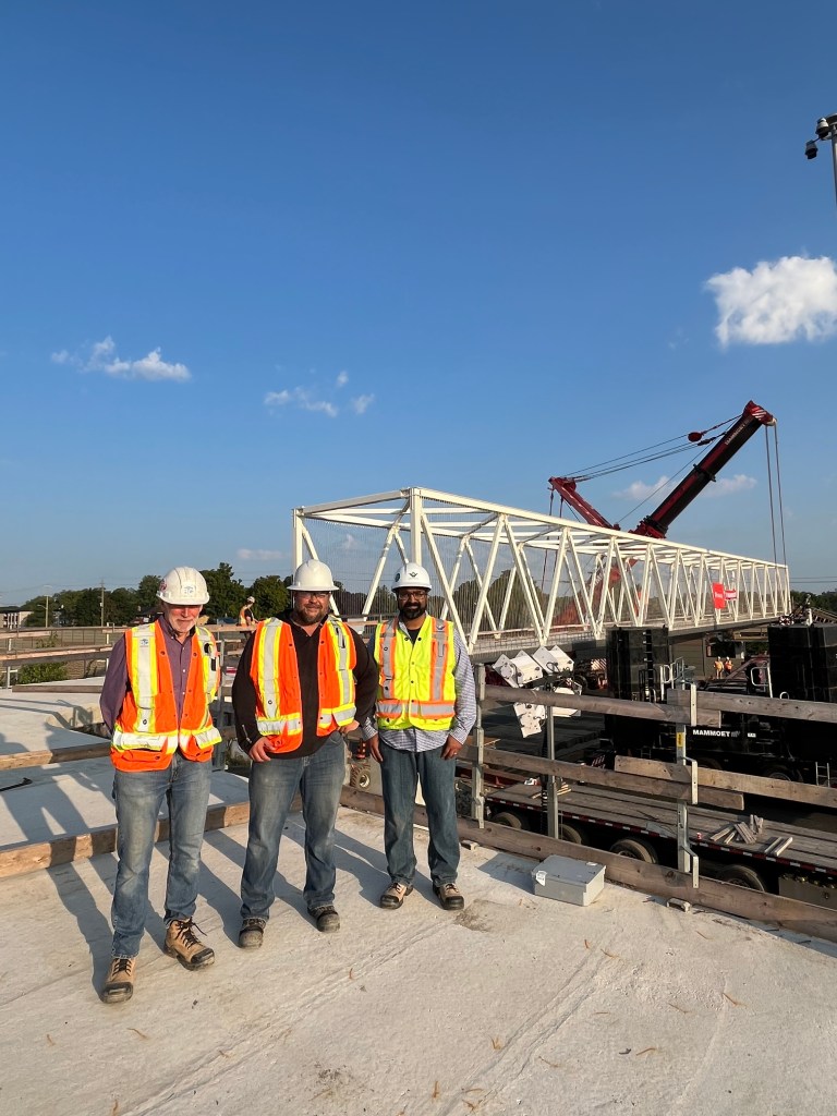 Three workers in orange vests stand in front of a new pedestrian bridge installed over Highway 7/8 in Kitchener.