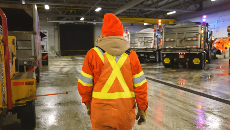 A woman wearing an orange high-visibility jumpsuit walks through a large facility past large machines.