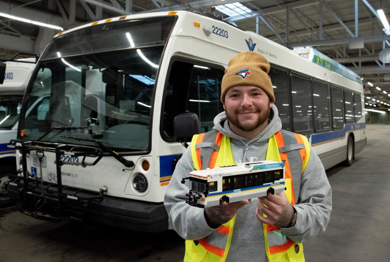 A man holding a small Lego model of a GRT bus stands in front of the full size vehicle.