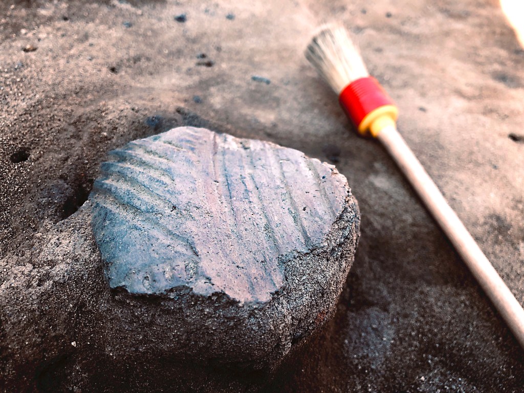 A ceramic piece in the dirt, where it was uncovered. A brush is beside it.