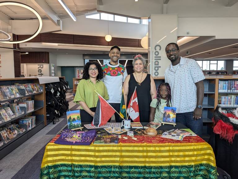 Five CCAWR volunteers of various ages stand smiling behind a booth with a colourful table cloth at a public library.