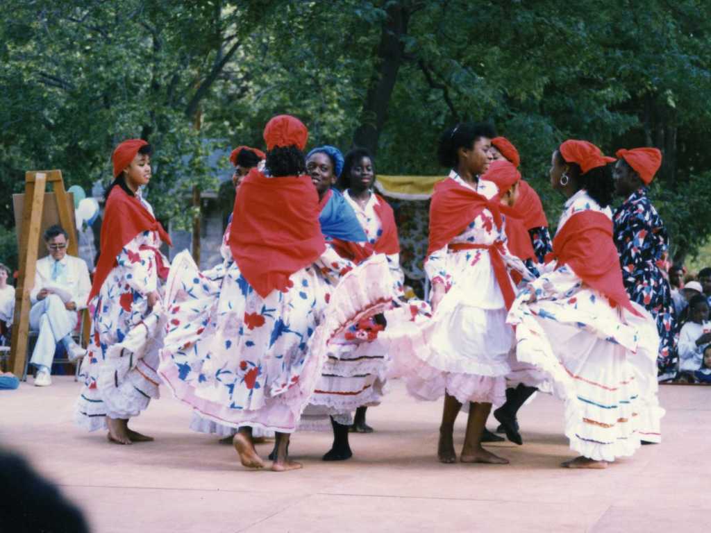 Young women in white and red dresses and red hats dance on a stage.