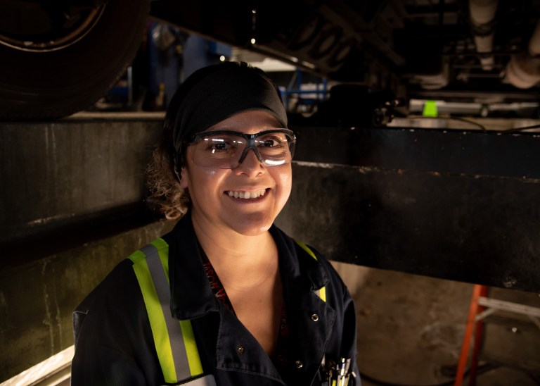 A woman wearing safety glasses and high-visibility clothes smiles in a dim garage.