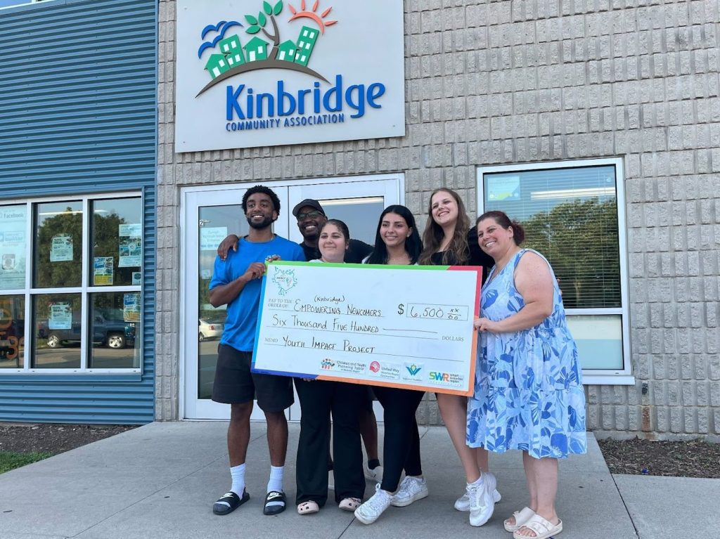 A group of six pose with a large cheque in front of the Kinbridge Community Association building.