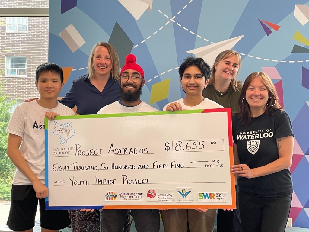 Young people smile while holding a large cheque in front of a colourful background.