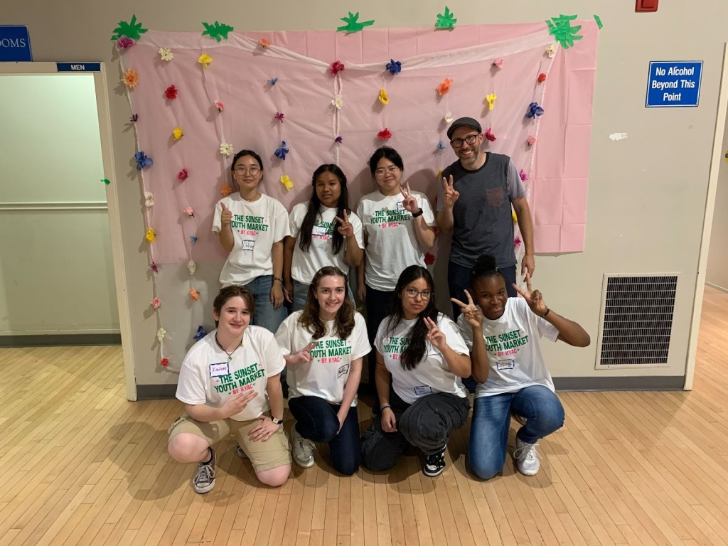 Young people in matching white t-shirts smile and gesture to the camera.