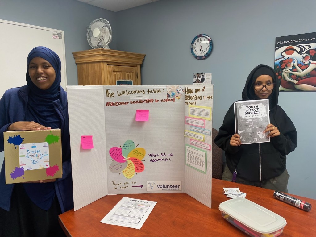 Two young women stand in front of a display board about their Youth Impact Project.