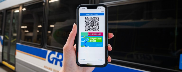 A hand holding a phone, showing the GRT Pay app. A bus is in the background.