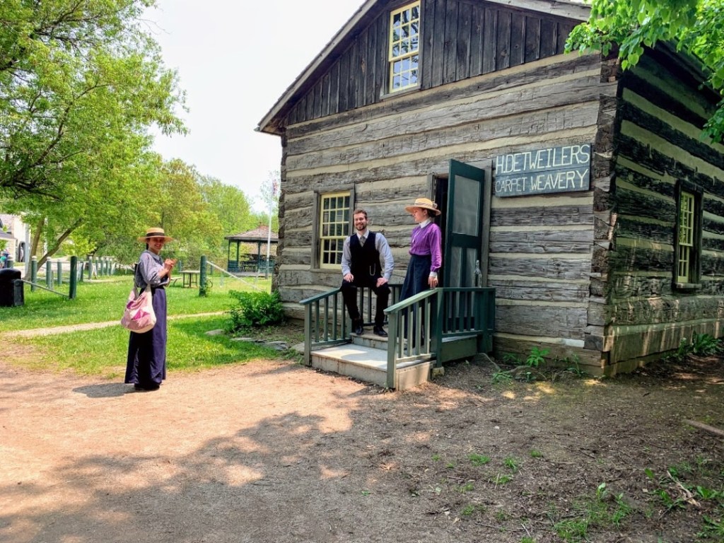 Staff in historical costumers stand outside an old-fashioned log structure at Doon Heritage Village.