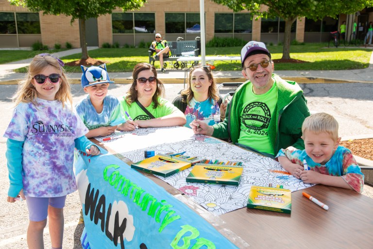 Adults and children sit around an outdoor table with paper and colouring materials.