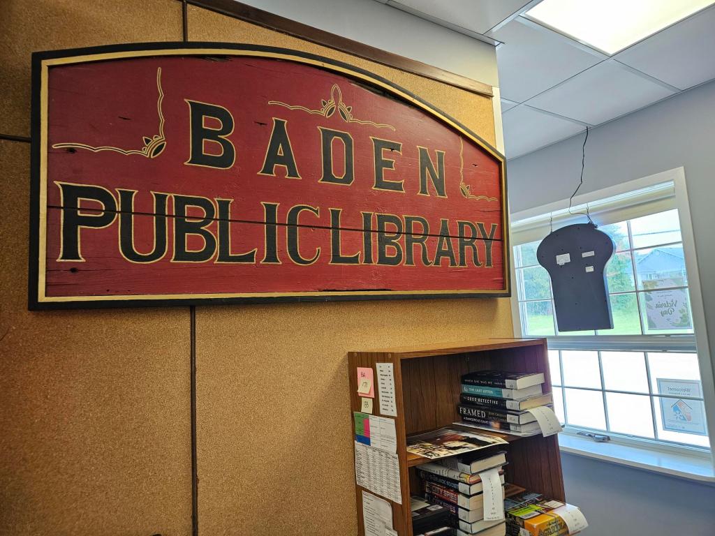 The restored, antique sign reading "Baden Public Library" mounted on a wall behind the service desk at the Baden library.