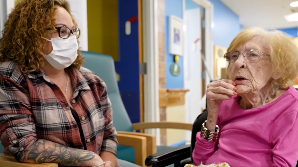 A younger woman wearing a medical mask sits with older woman in a care facility.