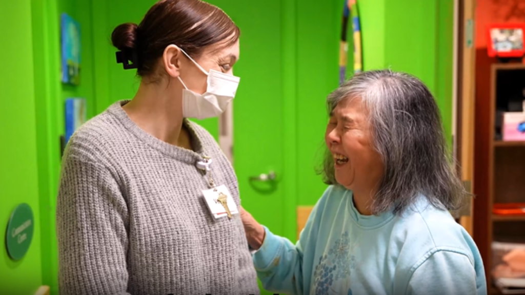 A younger woman wearing a medical mask stands with a laughing older woman.