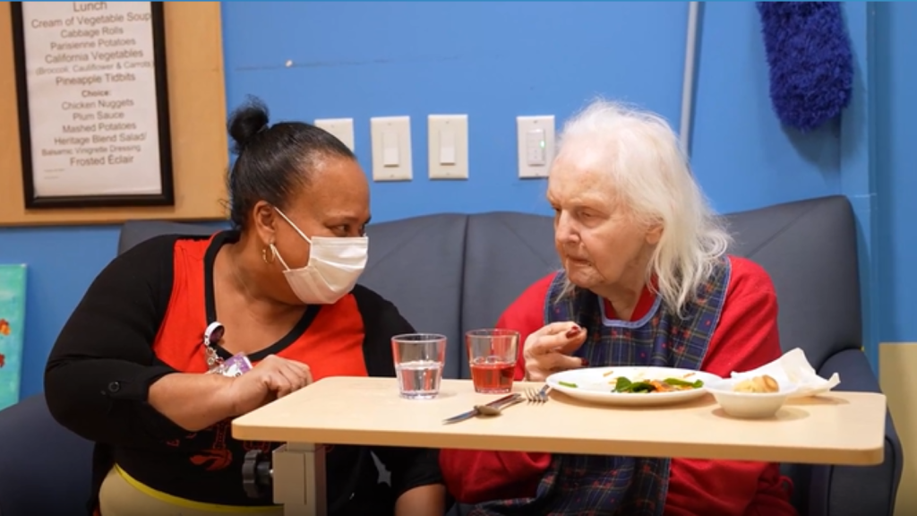 A younger woman wearing a medical mask sits with older woman at a table.